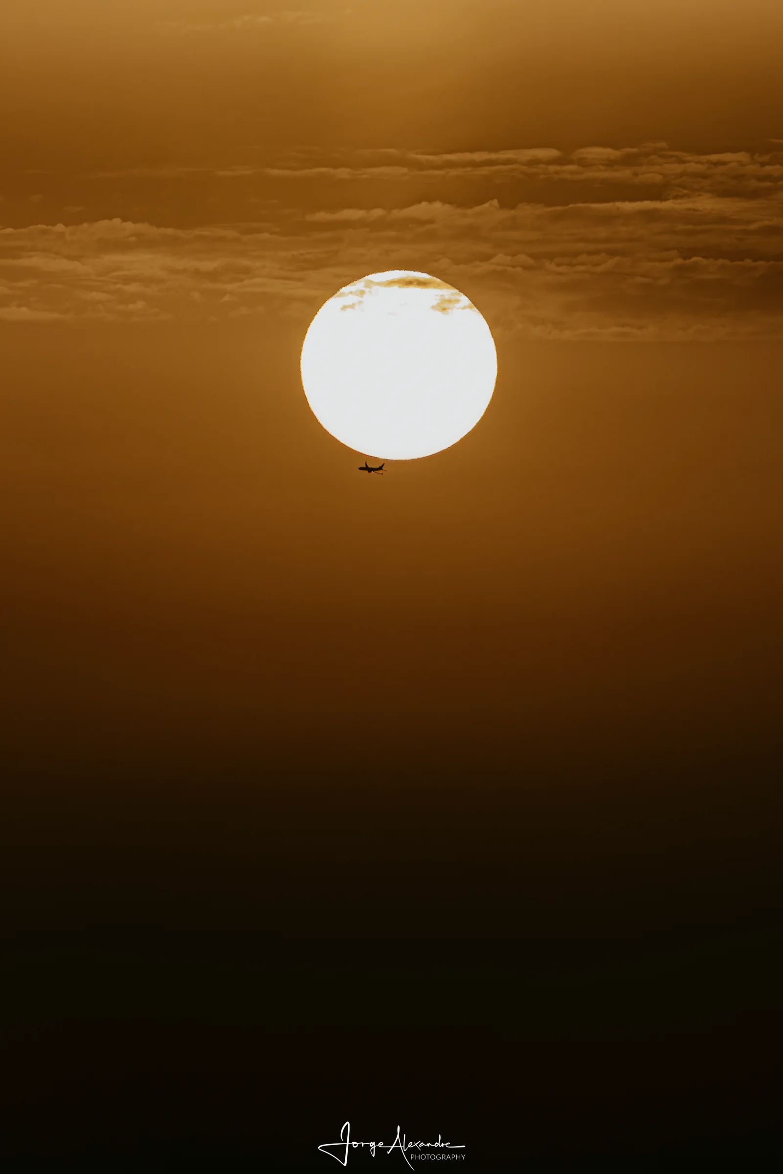 Tiny silhouette of an aircraft crossing below a large pale setting sun in amber haze.