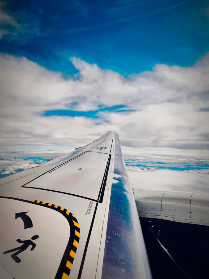 Airliner wing with walkway markings seen from the window in flight above a deck of white cloud and blue sky.