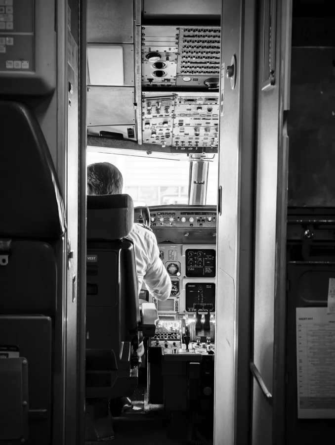 Black and white view through an open flight-deck door of a pilot from behind at the controls before pushback.