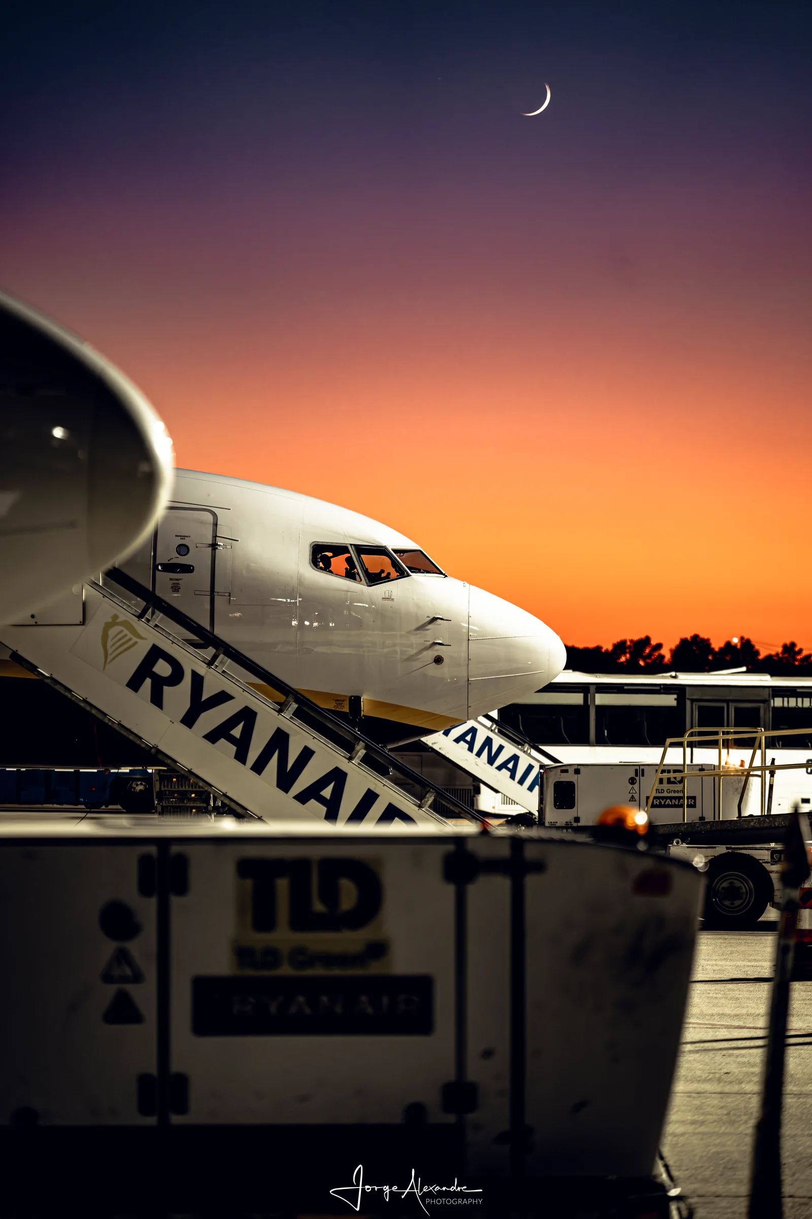 Nose of a Ryanair Boeing 737 with boarding stairs at dusk, under an orange and violet sky with a crescent moon.