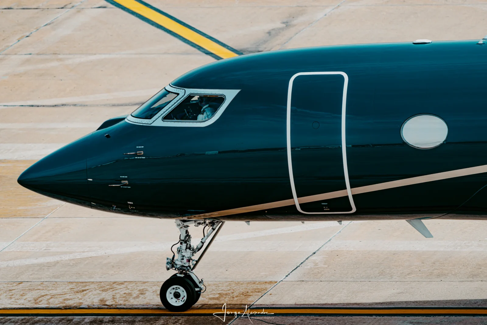 Glossy dark green business jet nose and cockpit taxiing over yellow lines on pale concrete.