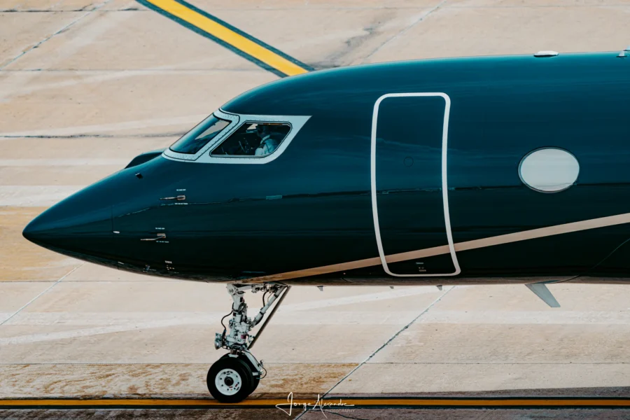 Glossy dark green business jet nose and cockpit taxiing over yellow lines on pale concrete.