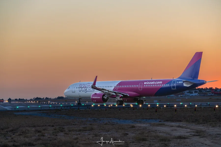 Wizz Air Airbus A321neo taxiing past illuminated runway lights under an orange sunset sky.