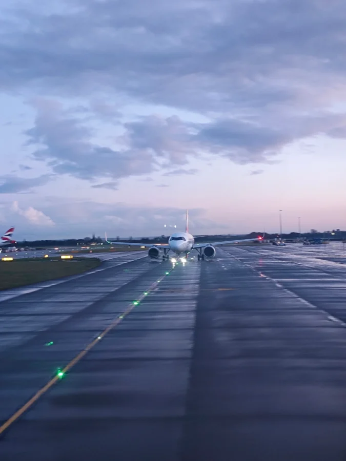 Airliner taxiing head-on along a wet taxiway at London Heathrow at dusk with green centreline lights.