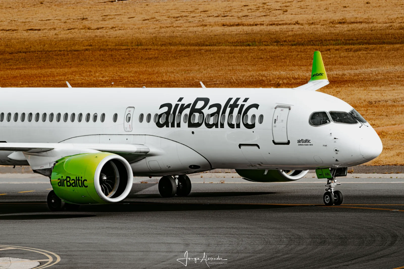 airBaltic Airbus A220-300 with lime-green engine and tail taxiing past golden grass.
