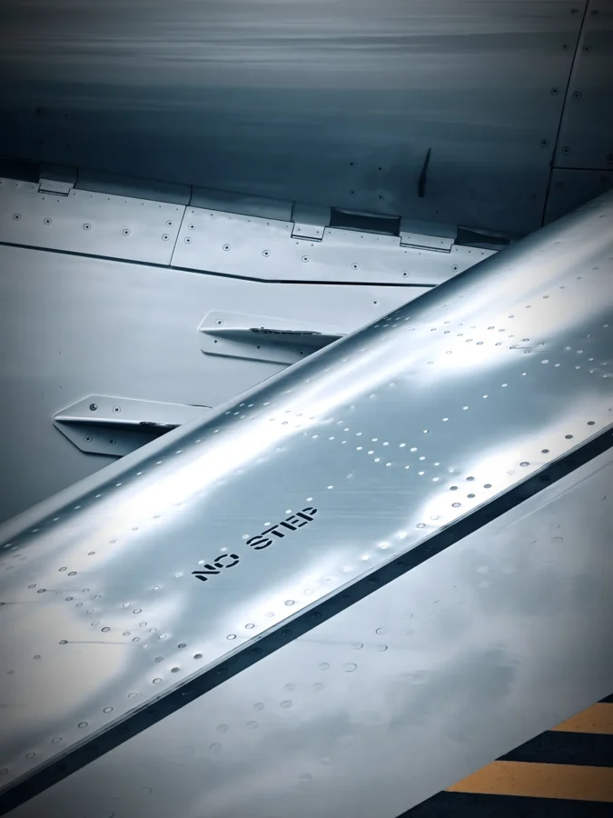 Close-up of an airliner wing surface with rivets, flap fairings and stencilled NO STEP lettering in cool blue tones.