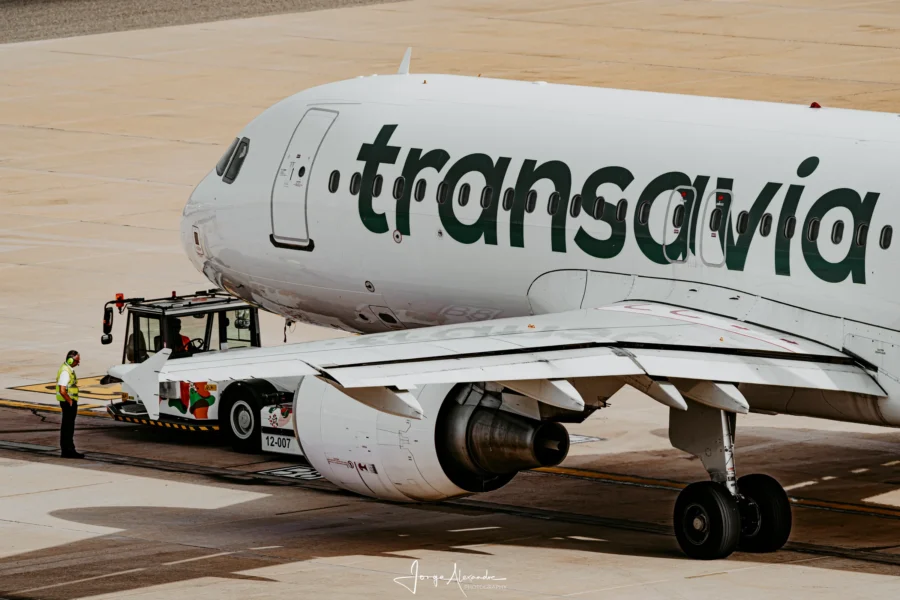 Transavia Airbus coupled to a pushback tug with a ground handler standing nearby.