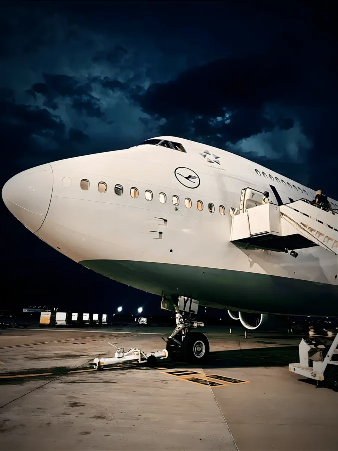 Nose of a Lufthansa Boeing 747-8 at night beneath stormy clouds, with boarding stairs at the forward door.