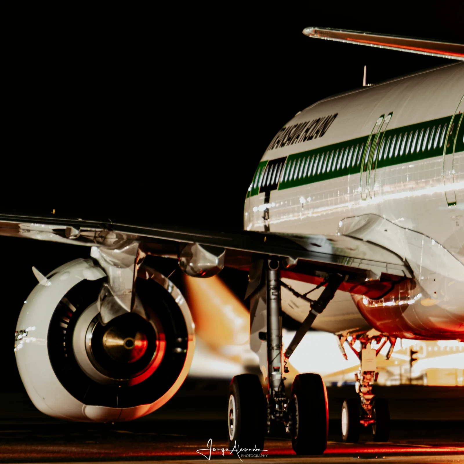 Night close-up of the Transavia Holland retro-livery Airbus engine, main gear and green-striped fuselage lit in warm light.