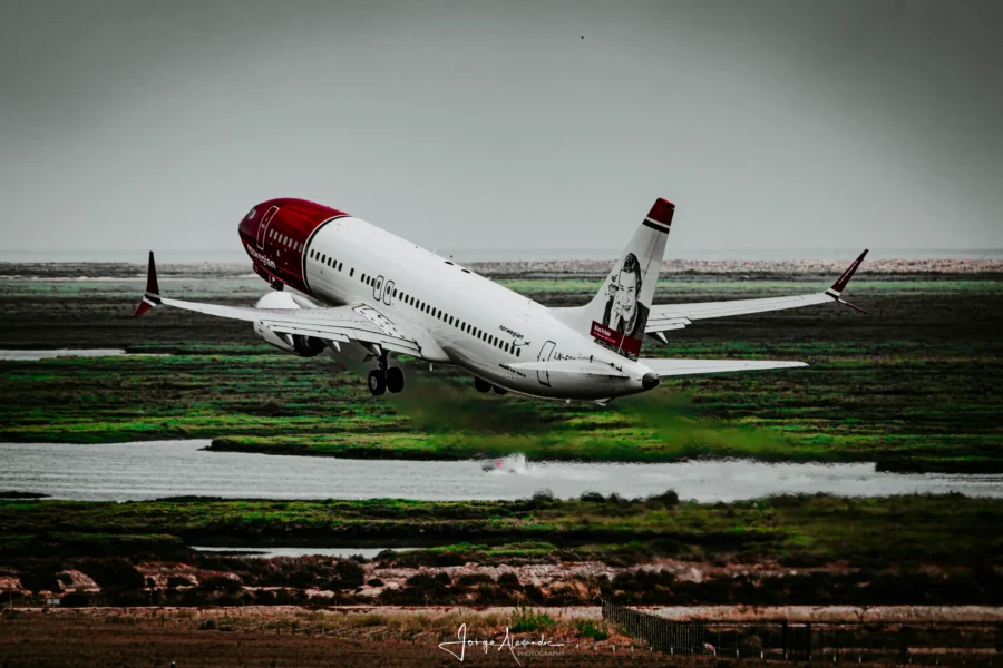 Norwegian Boeing 737 climbing after take-off above green salt marsh under a grey sky.