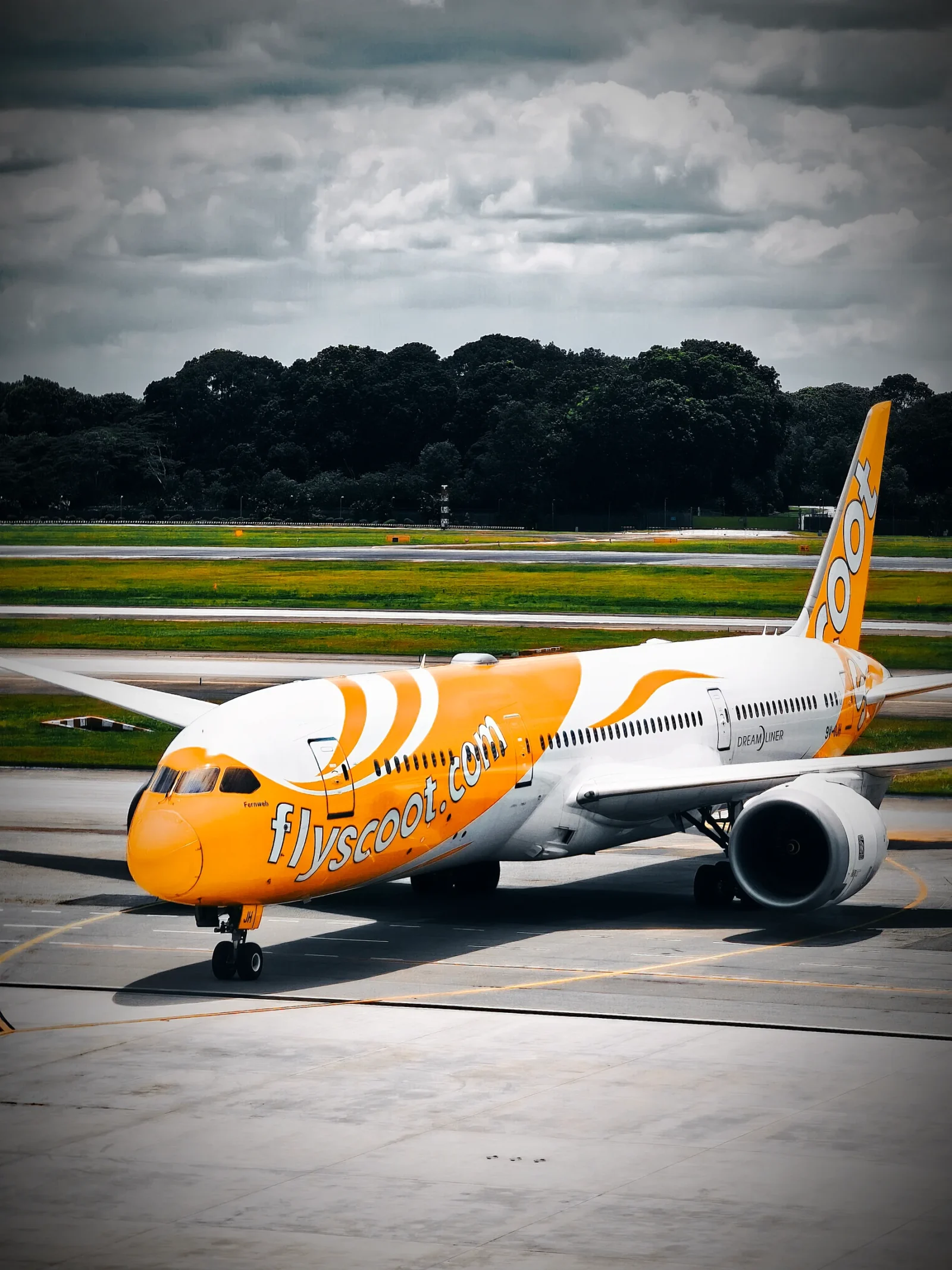 Scoot Boeing 787 Dreamliner in orange and yellow livery taxiing at Singapore Changi beneath a dark, cloudy sky.