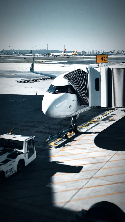 Airliner parked at stand 142 at Lisbon Airport, seen from above, with jet bridge, tug and strong diagonal shadows.