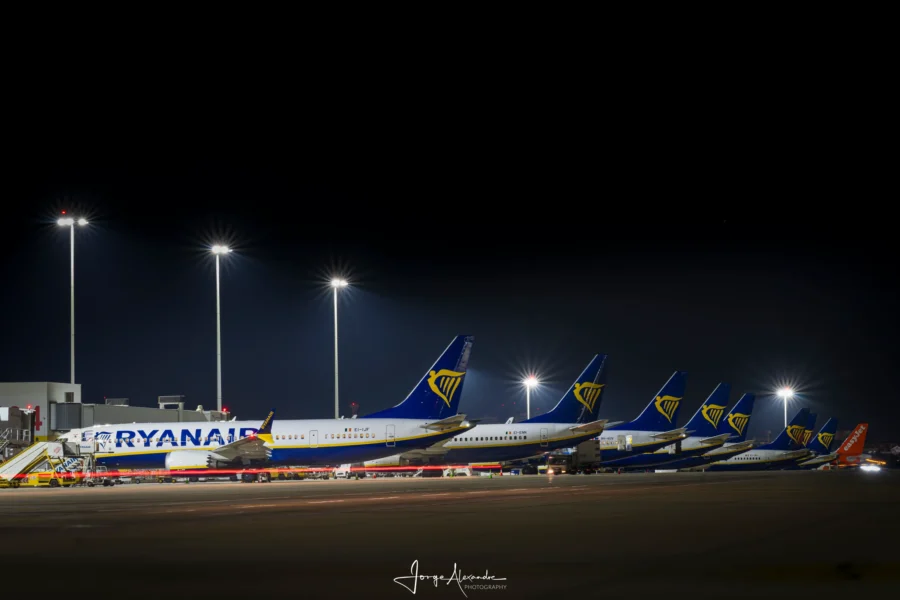 Line of Ryanair Boeing 737s with blue and gold tails parked along the terminal at night beneath floodlight masts.