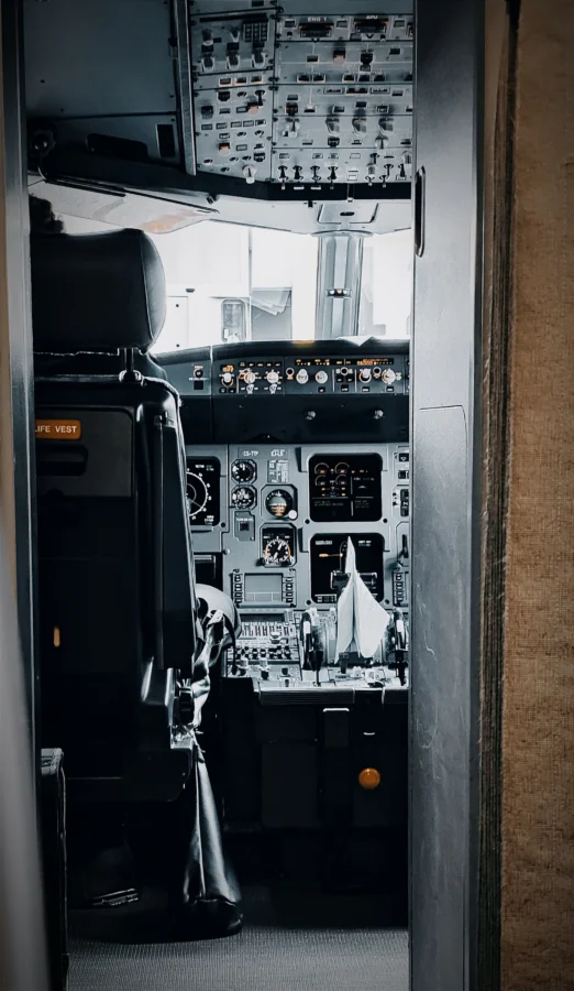 View through an open aircraft flight-deck door showing the captain's seat, overhead panel and instruments.