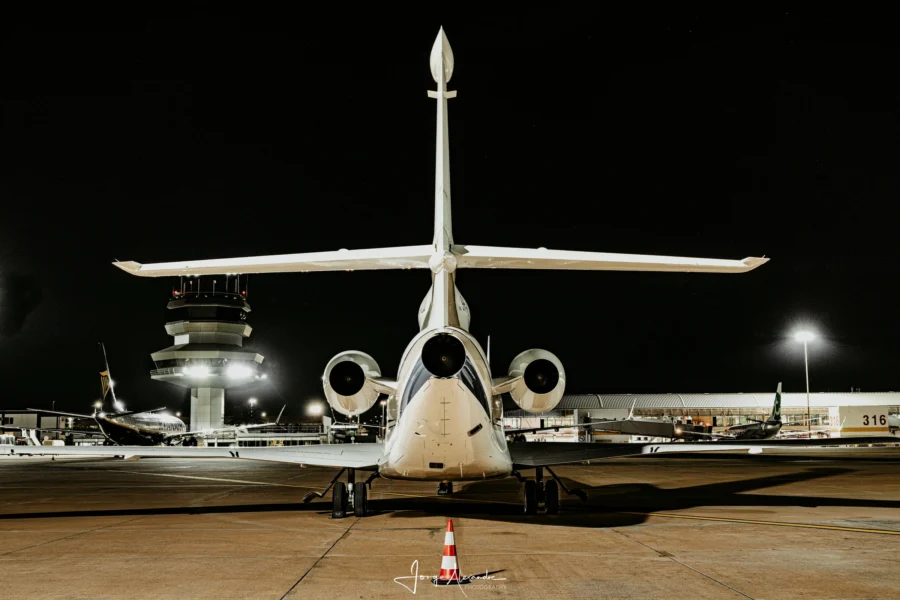 Dassault Falcon business jet seen from directly behind at night, with the lit airport control tower in the background.