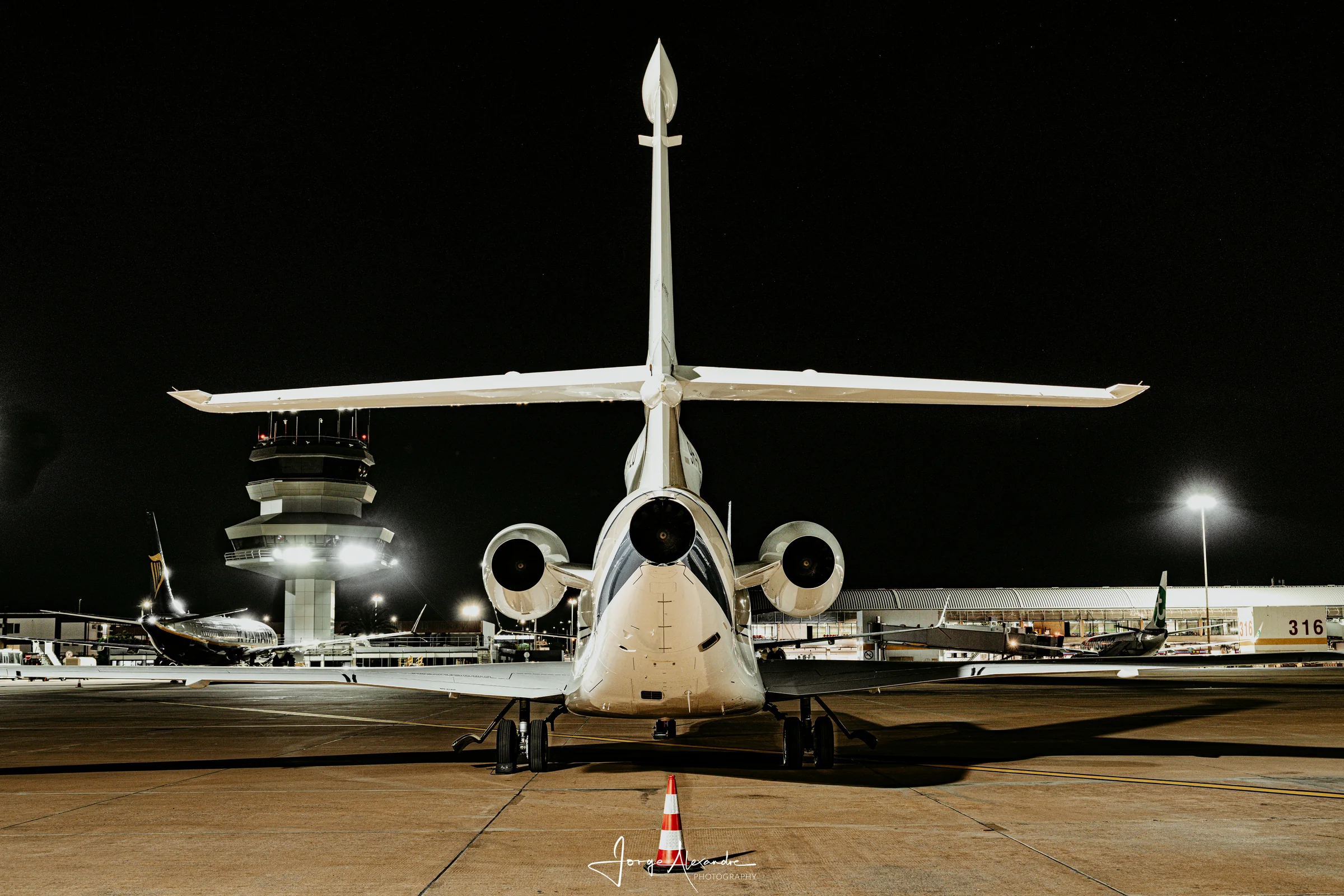 Dassault Falcon business jet seen from directly behind at night, with the lit airport control tower in the background.