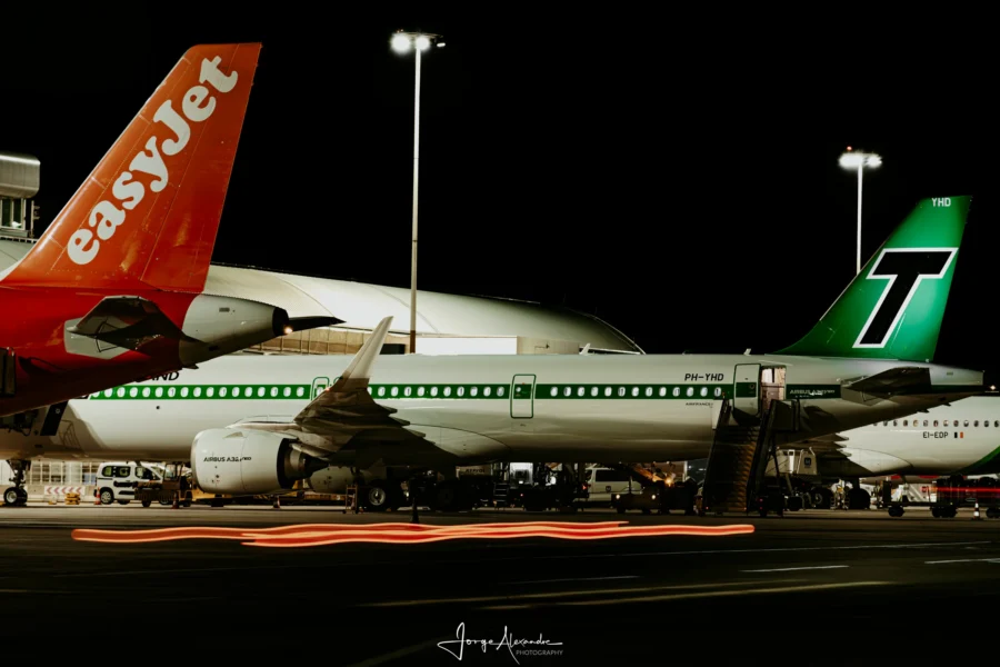 easyJet tail in the foreground and Transavia retro-livery A321neo at its stand at night, with red light trails across the apron.