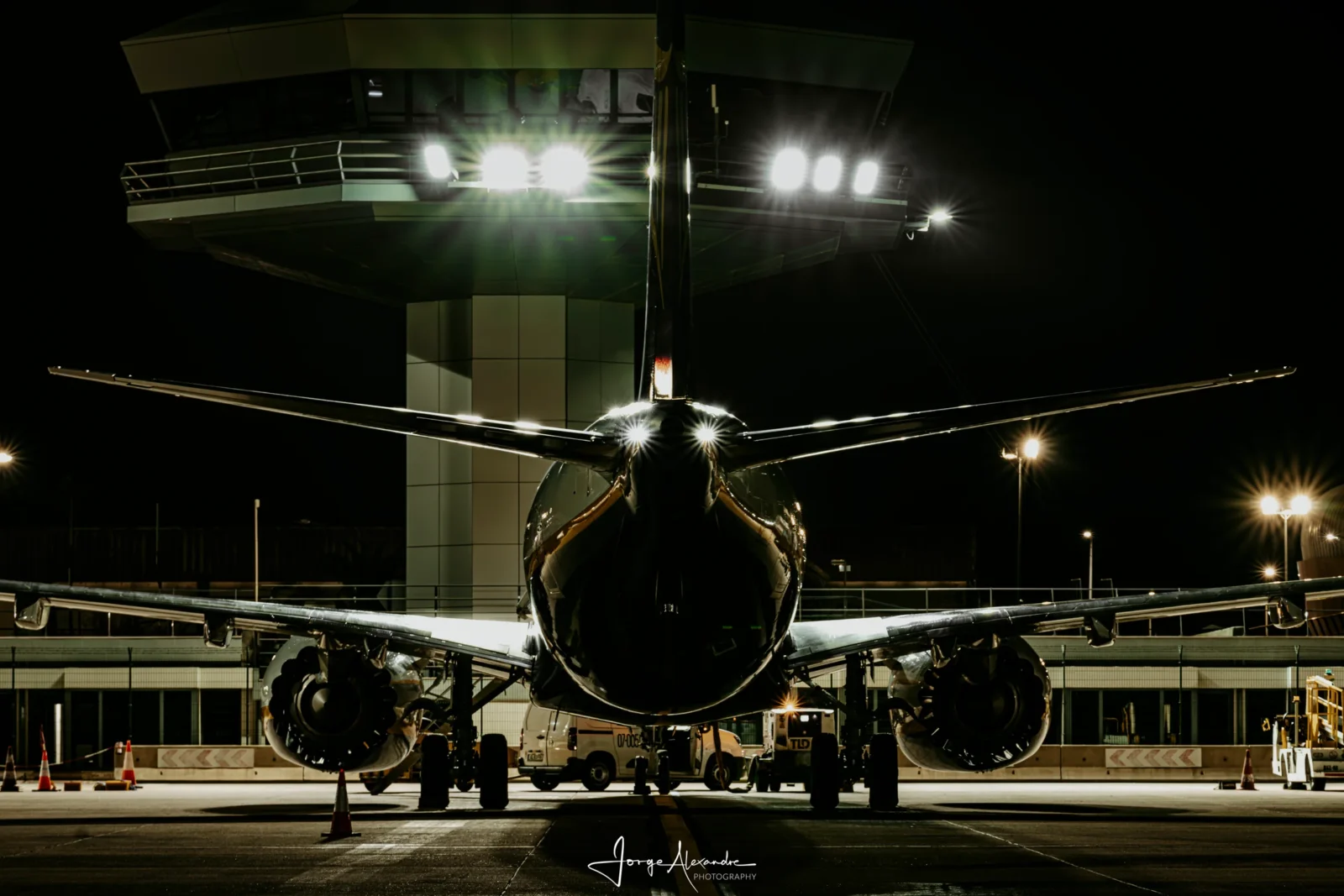 Airliner photographed from directly behind at night, silhouetted against the floodlit airport control tower.